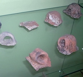 Na slici se vide komadići glinenih posuda neodređenih oblika. Predmeti stoje na staklenoj podlozi, odnosno posloženi su u vitrini.